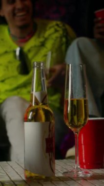 Bira şişesinin dikey yakın plan görüntüsü, bir bardak şampanya ve fayanslı masanın üstünde plastik bardak, arka planda arkadaşlarıyla eğlenen genç adam.