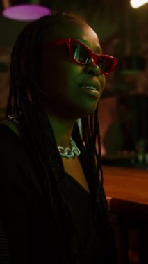 Gece kulübünde bar tezgahında otururken arkadaşlarıyla iletişim kuran Siyah Gen Z kızının dikey görüntüsü