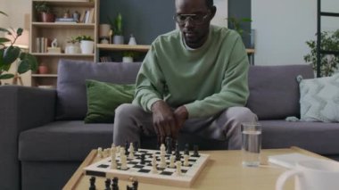 Afro-Amerikan bir adamın oturma odasında rahat bir koltukta oturup yeteneklerini geliştirmek için satranç taktikleri uyguladığı pozlar.