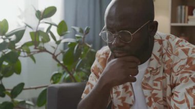 Gözlüklü ve rahat gömlekli düşünceli bir siyah adam, elini çenesinde tutuyor, evdeki antrenman sırasında satranç stratejisine konsantre oluyor.
