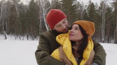Romantik çift kucaklaşıyor, hem gülümsüyor hem de fotoğraf için poz veriyor, karlı bir manzarada arka planda ağaçlardan oluşan bir ormanla dikiliyorlar.