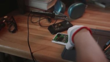 Bilinmeyen erkek teknisyenin, ahşap masadan kırık telefonu alıp, büyüteç yardımıyla ana kartı incelediği yakın çekim görüntüsü.