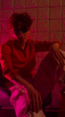 Kameraya yaslanmış ve neon ışıklı sandalyede otururken gülümseyen kıvırcık saçlı, gözlüklü Z geninin dikey portresi.