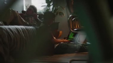 Genç müteahhit dizüstü bilgisayarda programlama yaparken yerde yatarken arkadaşının dinlendiği ve gitar çaldığı yere yaslanıyor.