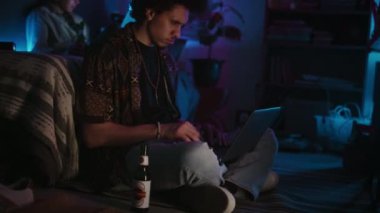 Bilgisayarlı, teknik sorunları çözen ve yerde neon ışıkla otururken bira içen odaklı Orta Doğulu genç müteahhit, arka planda kod yazan bir arkadaş.