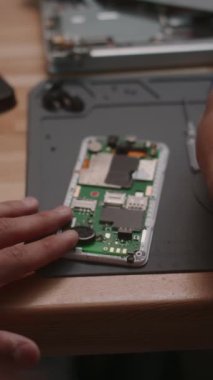 Ahşap masadaki bozuk akıllı telefonun mikroçiplerini tamir eden tanınmayan genç adamın dikey yakın çekimi.