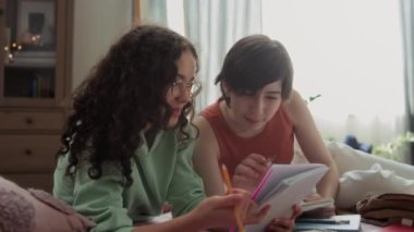 İki genç kız rahat bir odada birlikte ders çalışıyor, yatakta dinleniyor, kitabına dikkatle bakıyor ve ödevlerini tartışıyorlar.