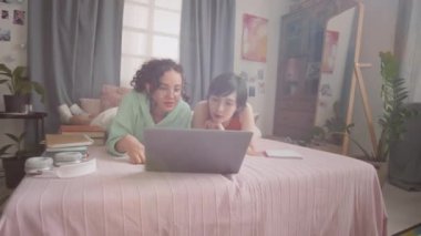 İki genç kızı yatakta yatarken, güneşli yatak odasında dinlenirken, gülerken ve dizüstü bilgisayar ekranına bakarken çek.