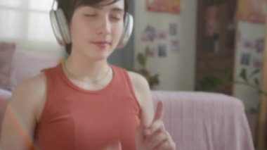 Genç bir kızın, rahat bir güneş ışığı odasında otururken, kulaklıkları açık müzik dinlerken, gözleri kapalıyken, ellerini ve başını çırpınırken yakın plan çekimleri.