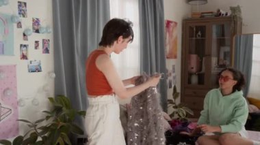 Işıltılı, güneşli yatak odasında giyinen iki genç kız, ışıl ışıl gümüş bluzunu deneyen bir kız, arkadaşı yatakta oturup onu izlerken...