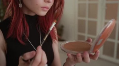 Kızıl saçlı genç bir kızın, gözlere ve buruna bronzlaştırıcı sürerken, kozmetik fırça kullanırken ve oturma odasında makyaj yaparken el aynasına bakarken,