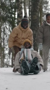 Vertical view of young man pushing his spouse who sliding on sled down snowy path, he and daughter running after woman