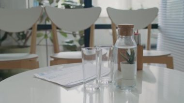 Modern hastanenin bekleme salonundaki yuvarlak beyaz masa üzerinde su şişesi, iki bardak, tıbbi belgeler ve işaret tutacağı yakın çekim görüntüsü.