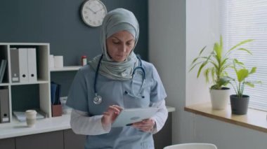Tıbbi önlüklü ve steteskop takan mavi tesettürlü kadın portresi, tıbbi ofiste dikilip dijital tablet kullanan, sonra da kameraya bakan...