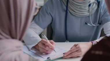 Kadın doktorun tesettürlü, hastalarını inceleyen, soru soran ve kağıda not alan bir fotoğrafı.