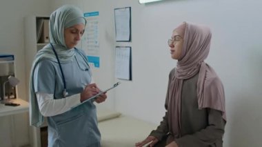 Hasta muayene koltuğunda oturup hastanedeki durumu hakkında konuşurken tesettürlü kadın doktor sağlık kontrolü yapıyor ve kağıda not alıyor.