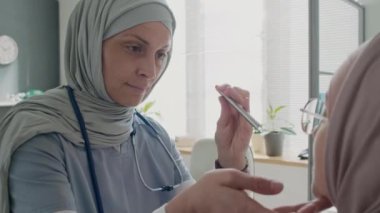 Kadın doktorun tesettürlü yakın plan fotoğrafı. Bir eli hastanede, elinde kalem ışığı olan Müslüman bir hastanın ağız boşluğunu kontrol ediyor.