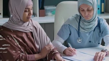 Hiyerarşik kadın doktor ve hasta portresi kameraya bakıyor, randevu sırasında masasında otururken nazikçe gülümsüyor.