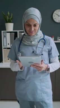 Klinikte iş günü dijital tablet kullanırken tesettür ve tıbbi önlüklerle gülümseyen ve kameraya bakan kadın doktorların dikey görüntüleri.