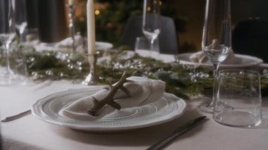 Beyaz tabaklar, boynuz tutacağıyla süslenmiş peçeteler, cam eşyalar, mumlar ve arka planda hafif ışıklandırılmış Noel ağacı olan ebedi yeşil süslemeler.