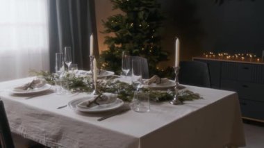 Gümüş tutacaklarda beyaz mumlar, her zaman yeşil olan süslemeler ve arka planda Noel ağacı ve sıcak ip ışıklarıyla dekore edilmiş rahat oturma odasında cam süslemeler.