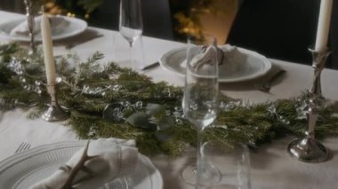 Çam dalları, okaliptüs ve cam boncuklarla çevrili masa örtüsü ve mumlarla çevrili ev yemeği masasındaki beyaz masa örtüsü.