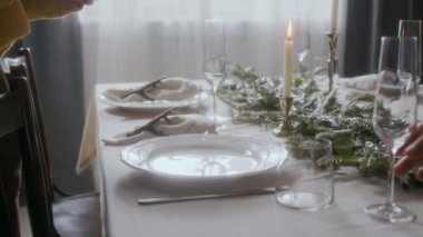 Beyaz tabak ve peçeteyi dikkatlice mumlarla, çam dallarıyla ve camlarla süslenmiş sofraya yerleştirirken evde Noel kutlaması için hazırlanan kadını yakından izleyin.