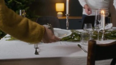 Noel masasına peçeteyle tabak koyan, çam dallarıyla süslenmiş, bir yandan da kocasının yardımıyla bayram yemeği hazırlayan tanınmamış kadının ellerini yakın çekim yapın.