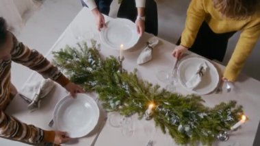 Yeşillik çelenk ve mumlarla süslenmiş sofraya düzgün sofra, çatal bıçak ve peçeteler yerleştiren üç arkadaşın yüksek açılı görüntüsü, evde Noel yemeği için hazırlanıyor.