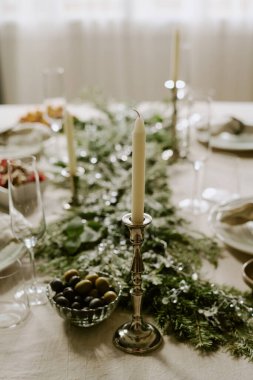Masadaki çam ağacından çelenk, kristal şampanya kadehleri ve tabaklar üzerindeki güzel gümüş mum tutacağında beyaz uzun mumların dikey açılı görüntüsü.