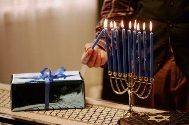 Medium close up of unrecognizable Jewish man burning candle with other one while standing next to table with candlestick, gift and Torah clipart