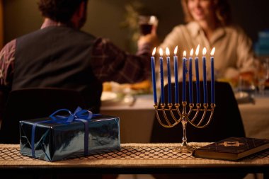 Medium close up of lit candles, gift and Torah on table with white tablecloth, unrecognizable Jewish couple sitting at table and drinking wine in blurred background clipart