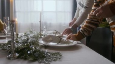Tanınmayacak haldeki dostların elleri, çatal bıçak takımı ve bardakları evde Noel için hazırlanırken yeşil çelenkler ve mumlarla süslenmiş şenlikli yemek masasına yerleştirirken yakın çekim.