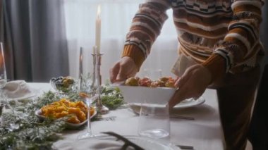 Örgü desenli süveter giyen bir adamın, evde Noel kutlaması için hazırlanırken, yeşil çelenkler ve mumlarla süslenmiş şenlikli yemek masasına meyve koyuşunun yakın görüntüsü.