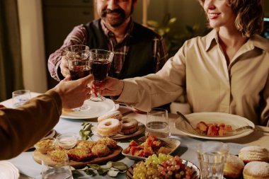 Medium close up of unrecognizable friends clinking crystal glasses with red wine while celebrating seasonal holiday at table with different types of treats clipart