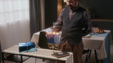 Kippah ışıklandırma menorah mumları takan Yahudi adam Hanuka kutlaması için rahat oturma odasında, arka planda şenlikli yemek masası olan