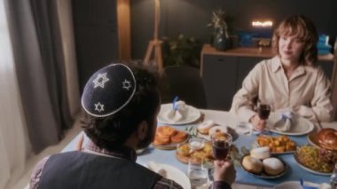 Kippah giymiş bir adam elinde şarap kadehi tutarken ve karısıyla konuşurken yemek masasında oturup evde geleneksel Yahudi yemeğinin tadını çıkarıyor.