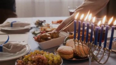 Hanukkah 'ı evde kutlamaya hazırlanırken menora mumlarıyla süslenmiş geleneksel Yahudi yemeklerini servis eden aile üyelerinin ellerini yakın.