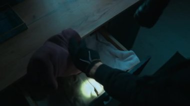 Erkek hırsızın el feneri kullanarak şifoniyer çekmecesini karıştırıp iki paket para bulup kameraya doğru eğildiğini görmek.