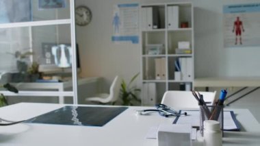 Masaları, posterleri, bilgisayarları, şeffaf ayırma duvarının arkasında göğüs röntgenlerini gösteren, rafları ciltlerle ve yerde saksı bitkileriyle dolu modern tıp bürosunun içi.