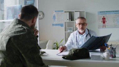Laboratuvar önlüklü orta yaşlı erkek doktor, modern tıp ofisinde masasında oturuyor ve MRI taramasını askeri üniformalı asker hastasına göstererek teşhisi açıklıyor.
