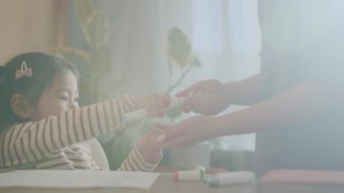 Çizgili kazak giymiş şirin küçük bir kız masanın başında oturmuş ve annesinden şapkasını çıkarmasına yardım etmesini istiyor, sonra pastel boyayı kadına uzatıp geri dönüyor.