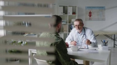 Laboratuvar önlüklü orta yaşlı erkek doktor askeri üniformalı asker üzerinde tansiyon monitörü kullanıyor ve tıbbi ofisindeki kağıt üzerinde notlar alıyor.