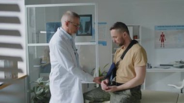 Laboratuvar önlüklü orta yaşlı erkek doktor. Askeri üniformalı askerin kırık kolunu muayene ediyor. Kolunda kol askısı var.