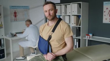 Askeri üniformalı yaralı askerin portresi, kırık kollu, hastanede muayene koltuğunda oturan, kameraya bakan, doktor arka planda tıbbi evrakları yazarken