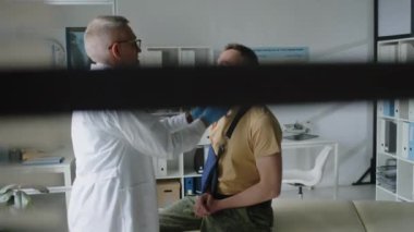 Laboratuvar önlüğü ve tıbbi eldivenlerle orta yaşlı erkek doktor. Askeri üniformalı askerin lenf düğümlerini okutuyor. Kolu kırık, klinikte muayene koltuğunda oturuyor.