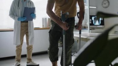 Askeri üniformalı tanınmayan bir asker koltuk değnekleriyle sağlık kontrolüne geliyor, bacağı kırık, kanepede oturuyor ve klinikteki doktor tarafından muayene ediliyor.