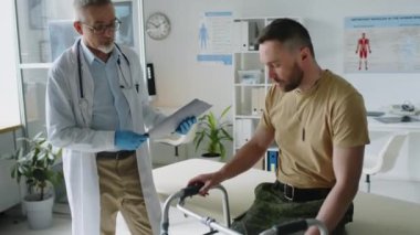 Orta yaşlı bir erkek doktorun, askeri üniformalı bir askerle bacak yaralanması hakkında röportaj yaparken, muayene koltuğunda otururken, kliniğe giderken yürüyüş çerçevesine yaslanırken,