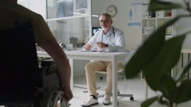 Laboratuvar önlüklü orta yaşlı erkek doktor, sağlık ofisinde masasında oturuyor, tekerlekli sandalyede emekli bir askeri randevu için görüyor.