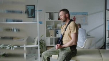 Kırık kolunu kaldıran yaralı askerin portresi, tıp ofisinde otururken, laboratuvar önlüklü doktor arka planda onun için tedavi öneriyor.